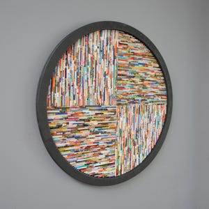 May include: A circular wall art piece made of rolled magazine paper in a variety of colours. The paper is arranged in a geometric pattern and framed in black.