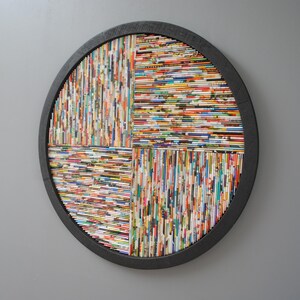May include: A round wall art piece made of rolled up magazine pages, arranged in a geometric pattern. The pages are in various colours and patterns, creating a colourful and textured effect. The art is framed in a black wooden circle.