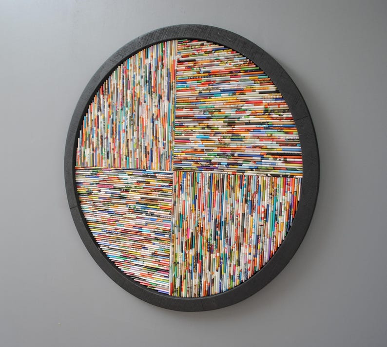 May include: A round wall art piece made of rolled up magazine pages, arranged in a geometric pattern. The pages are in various colours and patterns, creating a colourful and textured effect. The art is framed in a black wooden circle.