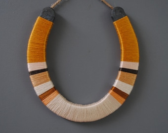 May include: A decorative horseshoe wrapped in yellow, cream, brown, and black yarn. The horseshoe is suspended by a natural twine loop, and the ends are capped with dark grey metal. This handcrafted item is a unique wall hanging.