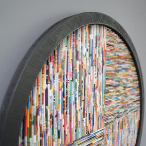 May include: A circular wall art piece made from colourful magazine strips, framed in a dark grey wood frame.