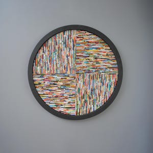 May include: A round wall art piece made of colourful magazine strips arranged in a geometric pattern. The strips are glued together to form a circular shape with a black frame.
