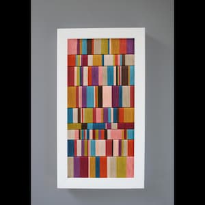 May include: A framed artwork featuring a vibrant, abstract design. The artwork is composed of vertical bands of various colors, including pink, blue, orange, and purple. The piece is encased in a white frame, creating a modern and colorful aesthetic.