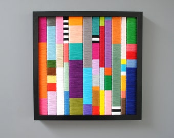 May include: A square wall art piece featuring vertical columns of tightly wrapped colourful yarn in a black frame. The composition consists of numerous multi-coloured segments including orange, pink, blue, green, purple, yellow, and black and white stripes.