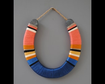 May include: A horseshoe wrapped in colorful yarn, with stripes of pink, orange, white, black, and blue. The horseshoe is hanging from a brown twine.