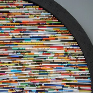 May include: A semi-circular wall art piece constructed from a range of colourful magazine pages. The pages are stacked and layered to create a distinctive and textured design. The piece is mounted on a black wooden frame.