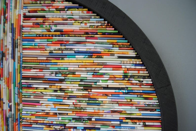 May include: A semi-circular wall art piece constructed from a range of colourful magazine pages. The pages are stacked and layered to create a distinctive and textured design. The piece is mounted on a black wooden frame.