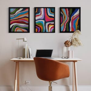 May include: Three framed abstract art prints with colorful, wavy lines and patterns. The prints are hung on a white wall above a desk with a laptop and a chair.