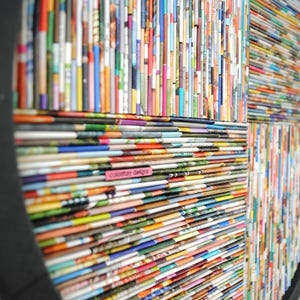 May include: A circular wall art piece made from rolled up magazine pages. The pages are arranged in a geometric pattern, creating a colourful and textured design. The piece is framed in a black circular frame.