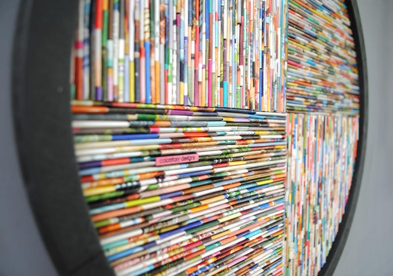 May include: A circular wall art piece made from rolled up magazine pages. The pages are arranged in a geometric pattern, creating a colourful and textured design. The piece is framed in a black circular frame.