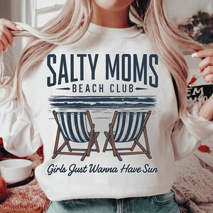 May include: White sweatshirt with a graphic design that says "Salty Moms Beach Club" with two beach chairs and the text "Girls Just Wanna Have Sun".
