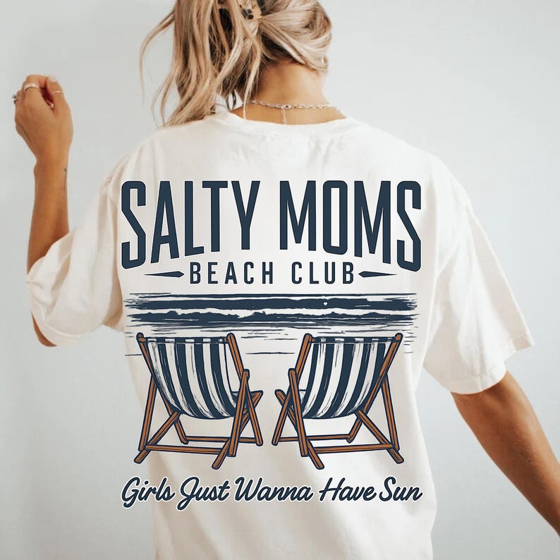 May include: White t-shirt with a graphic design that says "Salty Moms Beach Club" and features two beach chairs and the text "Girls Just Wanna Have Sun".
