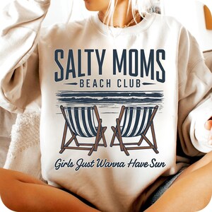May include: White sweatshirt with the text "Salty Moms Beach Club" and "Girls Just Wanna Have Sun" in black lettering. The graphic also features two beach chairs with blue and white stripes.