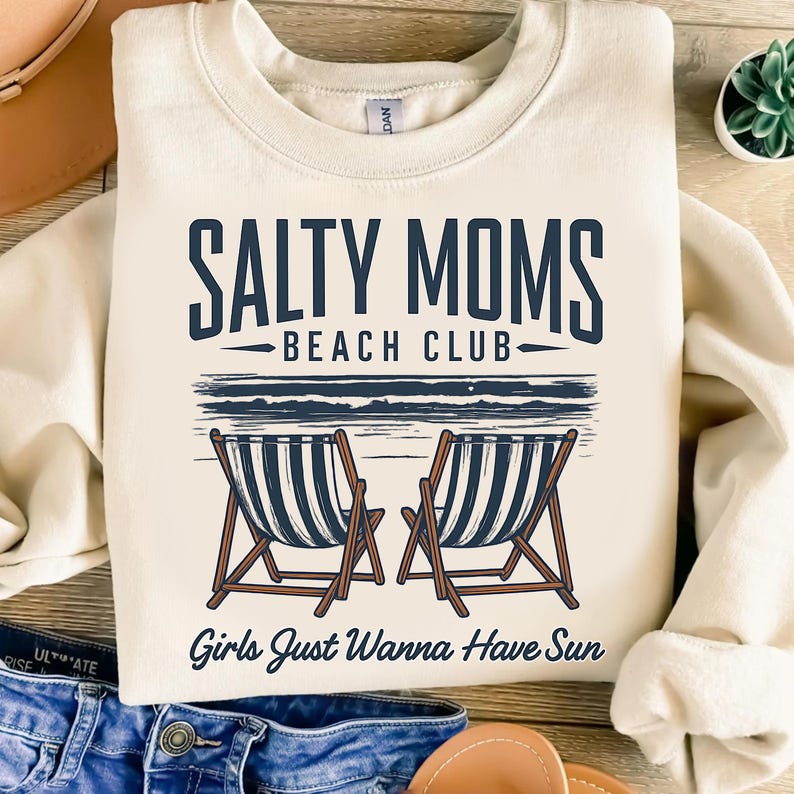 May include: A cream colored sweatshirt with the text "Salty Moms Beach Club" and "Girls Just Wanna Have Sun" in navy blue. The graphic features two beach chairs on a beach.