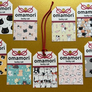 以下が含まれることがあります： 日本のお守り、おまもりラッキーキャットのコレクション。各タグにはユニークな猫のパターンがあり、「おまもりラッキーキャット」と書かれています。タグは赤い紐で繋がれています。