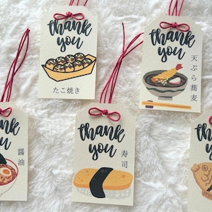 以下が含まれることがあります： 赤色の紐とリボンが付いた、クリーム色のギフトタグのコレクションです。「thank you」の文字が黒い筆記体で書かれており、たこ焼き、ラーメン、寿司、鯛焼きなど、日本の食べ物のカラフルなイラストが描かれています。