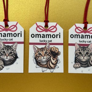 以下が含まれることがあります： 3つの日本のお守り、オマモリ。幸運の猫のイラストが描かれています。各タグには赤い紐と「omamori lucky cat」の文字があります。猫は様々なポーズで描かれています。