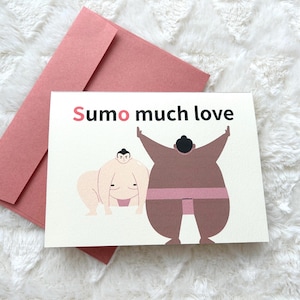 May include: A greeting card featuring the text "Sumo much love" and illustrations of two sumo wrestlers. The card rests on a pink envelope, set against a white, fluffy background.
