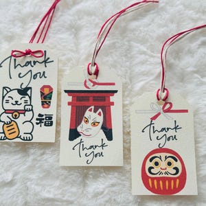 以下が含まれることがあります： 赤と白の紐が付いた、クリーム色のギフトタグ3枚。各タグには、招き猫、狐の面、ダルマ人形など、それぞれ異なる日本のイラストが描かれています。「Thank You」の文字が各タグに書かれています。