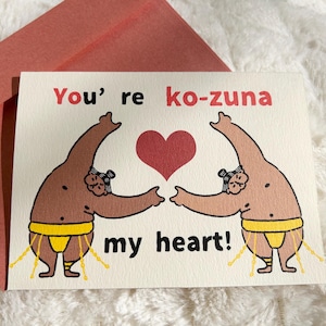 May include: Greeting card with a red envelope. The card features two cartoon sumo wrestlers reaching towards a red heart. Text on the card reads "You're ko-zuna my heart!" in red and black.