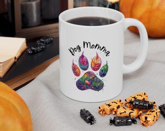 Könnte beinhalten: Weiße Keramiktasse mit den Worten "Dog Momma" und einem farbenfrohen Pfotenabdruck-Design. Der Pfotenabdruck zeigt Blätter im Aquarellstil in Blau-, Orange- und Grüntönen. Die Tasse ist mit einer dunklen Flüssigkeit gefüllt.