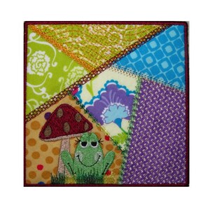 Può includere: Un quadrato di tessuto patchwork colorato con una rana verde, un fungo rosso e vari motivi floreali.