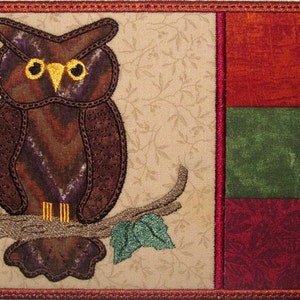 May include: A brown owl with yellow eyes is perched on a branch with a green leaf. The owl is against a beige background with a brown, green, and red patterned fabric to the right.