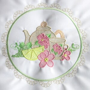 May include: Embroidered design of a teapot, teacup, lemon slice, and flowers in pink, yellow, and green colors. The design is surrounded by a green and gold lace border.
