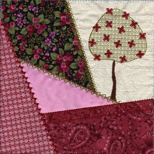 Può includere: Un blocco di trapunta patchwork con un design floreale rosa, rosso e verde. Il blocco presenta un albero stilizzato con un tronco marrone e foglie verdi.