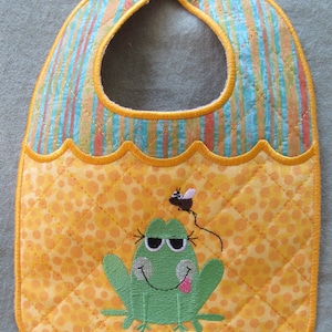 May include: A yellow and blue striped bib with a scalloped edge and a green frog with a fly on its head. The bib has a white button on the top.