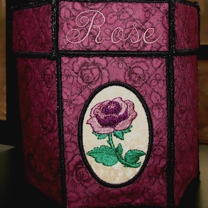 Peut inclure: Une boîte en tissu bordeaux avec une bordure noire et un motif de rose brodé. La boîte porte le mot "Rose" brodé sur le dessus.