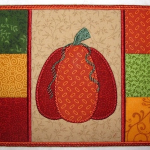 May include: A quilted patchwork design featuring a large orange pumpkin with a green stem, surrounded by squares of red, green, and yellow fabric with various patterns.