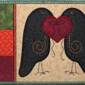 May include: Two black birds with gold stitching on their wings are facing each other with a red heart between them. The background is a cream color with a floral pattern.