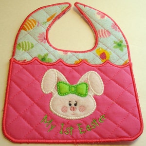 Op de afbeelding: Roze en blauwe baby-slabbetje met een gewatteerde roze zak met een geborduurde konijn met een groene strik en de tekst "My 1st Easter".