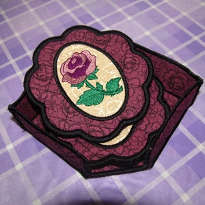 Può includere: Un porta tovaglioli in tessuto color borgogna con un bordo a festoni e un motivo floreale. Il motivo presenta una singola rosa con foglie verdi in una cornice ovale color crema.