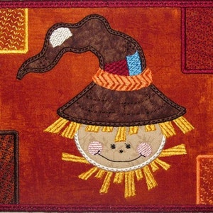 May include: A brown and yellow patchwork fabric scarecrow with a brown hat and a smiling face. The scarecrow is on a red background with brown and yellow squares.