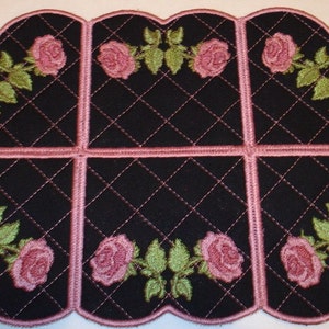 May include: A black quilted fabric coaster with a pink border and pink embroidered roses. The coaster is shaped like a heart with rounded edges.