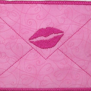 May include: A pink fabric envelope with a pink embroidered lip design. The envelope has a pink stitched border.