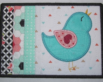 May include: A turquoise blue bird with a pink heart on its chest is embroidered on a white fabric background with a geometric pattern of small triangles. The bird has a yellow crown and a yellow beak. The fabric is surrounded by a black border.