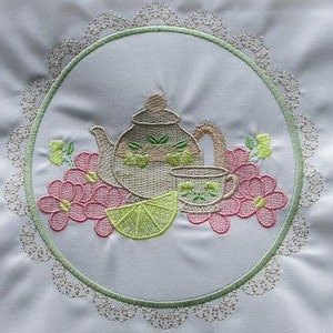 May include: Embroidered design of a teapot and teacup with pink flowers and green leaves on a white background. The design is surrounded by a green and white lace border.