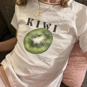 White Y2K aesthetic unisex t-shirt featuring a bold KIWI text, a vibrant kiwi graphic, and small retro-inspired text below. A tropical, playful vibe perfect for casual, streetwear looks