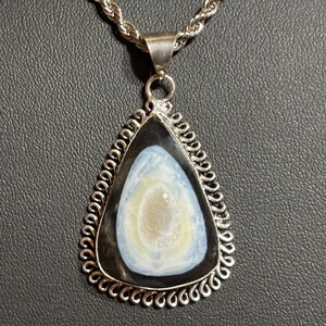May include: A silver necklace with a triangular pendant. The pendant features a black and white stone with a light blue and cream-colored center. The pendant is framed with a decorative silver border and hangs from a silver rope chain.