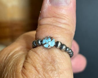 May include: A silver ring featuring a central turquoise stone with a light blue color and brown matrix. The ring is surrounded by small, round silver beads. The ring is shown on a person's thumb.