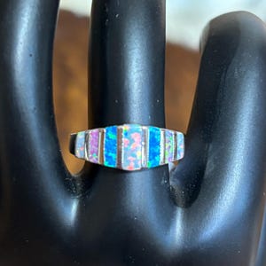 May include: A silver ring with a rectangular opal design. The opal has a mix of blue, pink, and white colors. The ring is set on a black surface.
