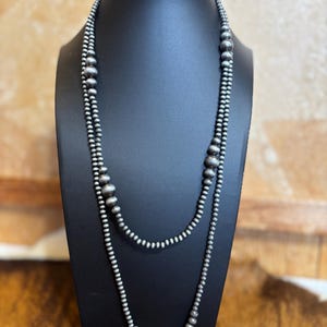 May include: A silver-toned necklace with two strands of varying bead sizes displayed on a black mannequin. The necklace features small, round beads and larger, textured beads, creating a layered effect. The beads have a matte finish.