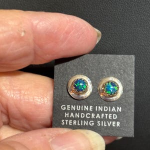 May include: A pair of sterling silver stud earrings with a round, bezel-set, iridescent gemstone. The earrings are displayed on a black card with the text "GENUINE INDIAN HANDCRAFTED STERLING SILVER".