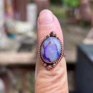 May include: A copper-toned ring with an oval, iridescent opal stone. The opal displays shades of blue, purple, and white. The ring has a twisted rope design around the stone and small star-shaped accents.