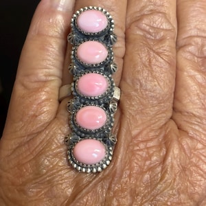 May include: A silver ring featuring five oval, light pink gemstones. The gemstones are set in individual bezels with a decorative border and are connected by a silver band. The ring is a statement piece of jewelry.