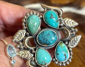 May include: A silver ring featuring a large central oval turquoise stone surrounded by four smaller pear-shaped turquoise stones. The setting includes decorative silver leaves and crescent moon shapes with beaded accents. A separate thin silver band with two small blue stones is also visible.
