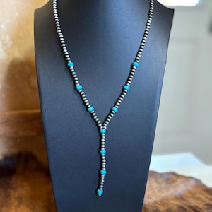 May include: A silver-toned necklace with turquoise-colored beads. The necklace is displayed on a black mannequin. The beads are small and round, with the turquoise beads spaced along the necklace's length. The necklace has a Y-shaped design.